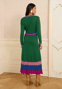 Rochie verde tricotată cu mâneci lungi, având un model zigzag multicolor în partea de jos și un guler cu dungi. Este purtată cu cizme cu toc de culoare cărămizie deschisă.