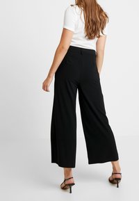 Zwarte wijde broek gemaakt van een gladde stof. Voorzien van een hoge taille, zijzakken en een getailleerde pasvorm, gestyled met zwarte hakken met banden.