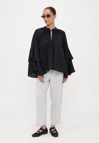 Black oversized blouse with layered bell sleeves, lightweight fabric, round neckline, and a front keyhole opening. Paired with light gray trousers.