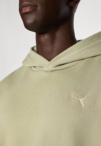 Sweat à capuche vert avec une capuche texturée et ajustable, et un petit logo Puma rose pâle sur la poitrine. Tissu lisse avec une coupe décontractée.