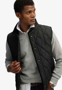 Chaleco negro acolchado con botones a presión, usado sobre un suéter gris. Presenta un cuello alto y costuras visibles. Fondo claro.