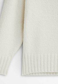 Pull en tricot beige clair avec une finition texturée et douce. Il présente un ourlet et des poignets côtelés pour une touche de détail. Design simple et épuré.