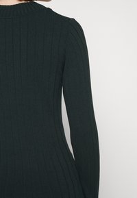 Gros plan sur une personne portant un haut à manches longues ajusté de couleur foncée à côtes, montrant le haut du dos et le bras droit sur un fond uni.