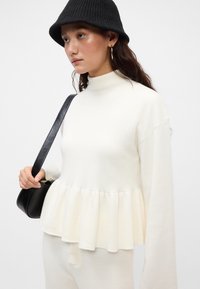 Maglione bianco a maglia con maniche lunghe e alto colletto; presenta un orlo a peplum. La modella porta una borsa nera e indossa un cappello a secchiello a coste nera.