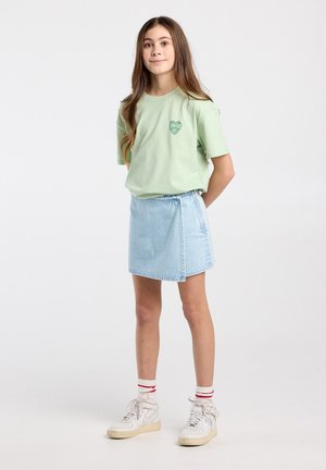Fille debout, les mains dans le dos, portant un t-shirt vert clair, une jupe en jean bleu clair, des baskets blanches et des chaussettes blanches à rayures rouges.