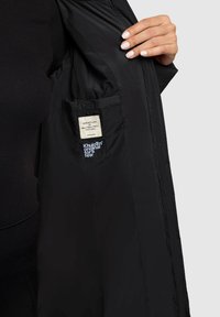 Unutrašnjost crne jakne prikazuje glatku satenastu teksturu, s etiketom koja sadrži bijeli tekst. Uključuje džep i vidljiv šav.