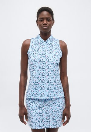 Γυναίκα που φοράει αμάνικο ανοιχτό μπλε τοπ με γιακά και ταιριαστή φούστα με μικρά σκούρα και ροζ φύλλα, στέκεται μπροστά σε λευκό φόντο.