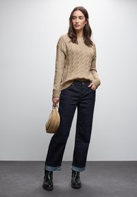 Maglione beige a trecce, jeans a gamba larga blu scuro con risvolti, stivaletti neri alla caviglia e una borsa rotonda marrone chiaro. Design semplice e testurizzato.