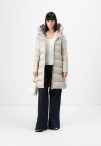 Licht beige pufferjack met capuchon, met doorgestikte secties en een ritssluiting aan de voorkant. Draagt een witte top en donkere flare jeans. Houdt een witte tas vast.
