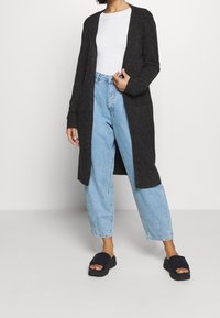 Lång grå cardigan i ett strukturerat tyg, med öppen framsida. Matchas med ljusblå högmidjade jeans och svarta sandaler.