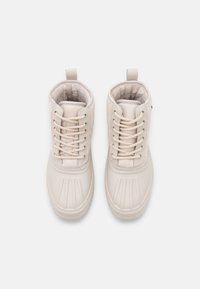 Vaaleanbeige nilkkurit, jotka on valmistettu synteettisestä materiaalista. Niissä on teksturoitu päällinen, nauhakiinnitys ja vetotappi takana.