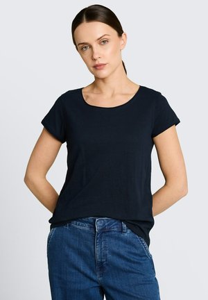 Femme aux cheveux foncés attachés en queue de cheval basse, portant un t-shirt noir uni à manches courtes et un jean bleu, debout les mains derrière le dos.