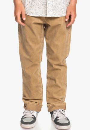 Cordhosen in Hellbraun mit geripptem Muster, versehen mit umgeschlagenen Bündchen, kombiniert mit grauen und weißen Sneakers.