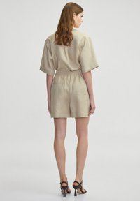 Combinaison en lin beige à manches courtes avec taille élastique, poches arrière et shorts ajustés ; complétée par des talons noirs à lanières.