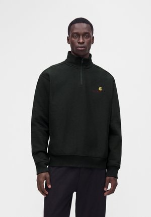 Man draagt een zwarte trui met kwart-ritssluiting en een klein geel Carhartt-logo op de borst en donkere broek tegen een witte achtergrond.