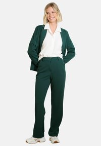 Completo verde realizzato in tessuto liscio, composto da un blazer e pantaloni coordinati. Sotto una camicia bianca con colletto. Modello con sneakers bianche.
