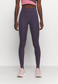 Fioletowe legginsy z wysokim stanem, wykonane z elastycznej tkaniny, charakteryzujące się gładką powierzchnią i minimalną ilością szwów. W połączeniu z jasnoróżowymi butami sportowymi.