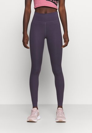Leggings morados de cintura alta, elaborados con un tejido elástico, que cuentan con una textura suave y costuras mínimas. Combinados con zapatillas deportivas de color rosa claro.