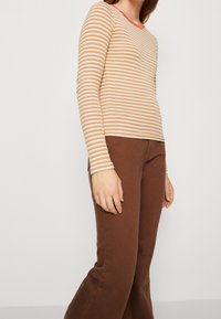 Langärmliges gestreiftes Oberteil mit einem Muster aus Creme, Orange und Blau, kombiniert mit braunen, eng anliegenden Hosen. Das Oberteil hat einen einfarbigen roten Ausschnitt.