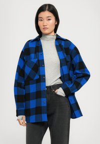 Camicia di flanella a quadri blu e neri con design a bottoni, taschino sul petto e vestibilità rilassata, indossata sopra un dolcevita grigio e jeans neri.