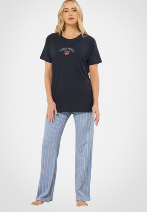 Kvinde iført sort "Sunday Brunch Club" T-shirt og lyseblå stribede bukser, stående barfodet mod en ensfarvet baggrund.