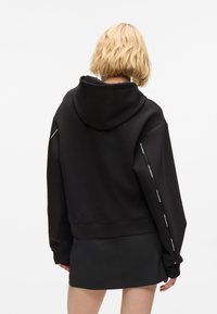Pessoa loira vestindo um hoodie preto com riscas de logotipo nas mangas e uma saia preta curta, vista de costas.