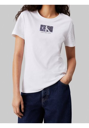 Frau trägt weißes Calvin Klein Jeans T-Shirt mit schwarzem Logo und dunkelblaue Jeans, eine Hand in der Tasche, vor grauem Hintergrund.