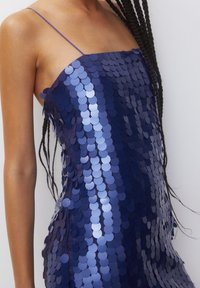 Robe bleu marine ornée de sequins circulaires superposés, avec des bretelles fines et une silhouette ajustée. Texture lisse et accents scintillants.