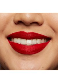 Röd matt läppstift täcker hela läpparna och framhäver en jämn och slät applicering med blanka vita tänder synliga. Huden har en naturlig ton.