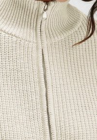 Nahaufnahme eines weißen gestrickten Pullovers mit geripptem Kragen und silbernem Reißverschluss in der Mitte, getragen von einer Person mit sichtbarem dunklem Haarstrang.
