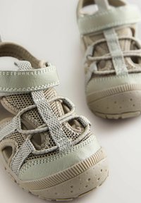 Beige och ljusgröna sandaler har en texturerad ovandel med mesh- och lädermaterial. Designen inkluderar justerbara remmar och en gummisula.