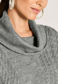 Frau mit großen silbernen Creolen und einem grauen Strickpullover mit weitem Kragen.