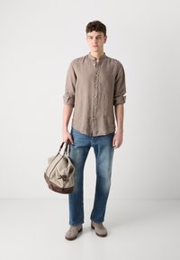 Linnen button-up shirt in lichtbruin, opgerolde mouwen, gecombineerd met blauwe jeans en grijze suède schoenen. Houdt een beige en bruine sporttas vast.