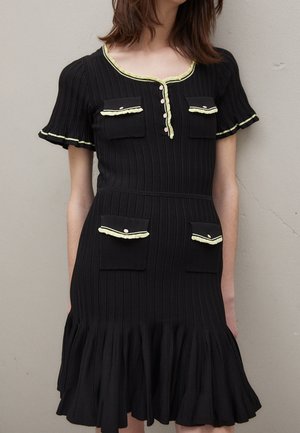 Schwarzes, geripptes Kleid mit kurzen Ärmeln, vier Klappentaschen und blassgelbem Rüschenbesatz am Ausschnitt, an den Manschetten und an den Taschenklappen.