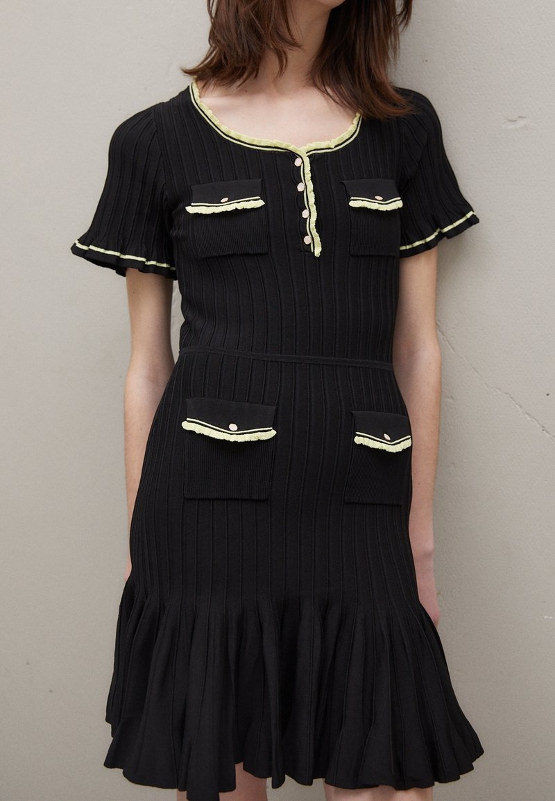 Robe noire côtelée à manches courtes, quatre poches à rabat et finition volantée jaune pâle sur l'encolure, les poignets et les rabats des poches.