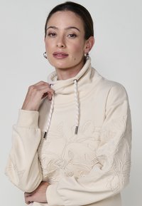 Sweatshirt beige avec un col haut, broderie florale détaillée sur les manches, et cordons torsadés avec des embouts métalliques. Texture douce.