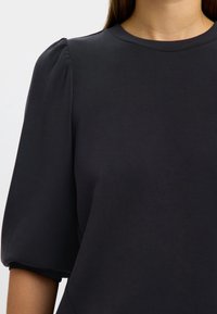 Melna īsām piedurknēm krekls ar noapaļotu kakla izgriezumu un pūkainām plecu detaļām. Gluda auduma ar smalku spīdumu, pieguloša silueta.