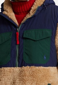 Giacca foderata in pelliccia con parte superiore blu navy, tasche flappate verdi e sezione inferiore in pelliccia marrone chiaro. Presenta un tirazip rosso e un cappuccio.
