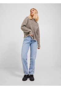 Strikket beige og brunstripete genser med høy hals, ribbet tekstur og kontrasterende kremfargede detaljer, sammen med lyseblå jeans og svarte sko.