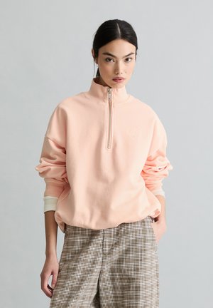 Frau mit dunklen Haaren im niedrigen Pferdeschwanz, die einen hellrosa Pullover mit Viertel-Reißverschluss und braune karierten Hosen vor neutralem Hintergrund trägt.