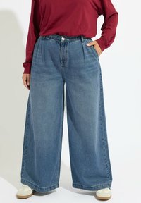 Niebieskie dżinsowe spodnie o szerokim kroju z wysokim stanem, plisami z przodu i kieszeniami; w połączeniu z kremowymi sneakerami i bordowym topem z długim rękawem.