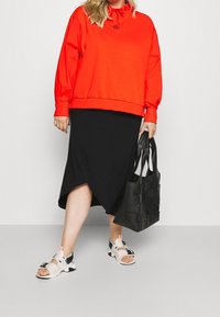 Červená oversized mikina s kapucí a šňůrkou, černá zavinovací sukně, černá tkaná taška přes rameno a bílé sandály s pásky a sponami.