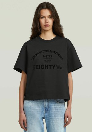 Vrouw met lang bruin haar, gekleed in een zwart oversized T-shirt met de tekst "G-STAR" en lichtblauwe jeans, staand tegen een effen achtergrond.