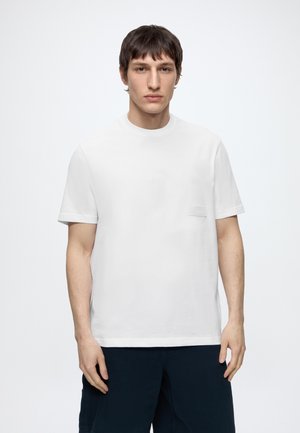 Jeune homme aux cheveux courts et foncés portant un t-shirt blanc uni à manches courtes et un pantalon foncé, debout devant un fond blanc.