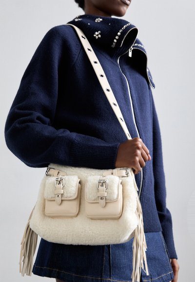 Άτομο που κρατάει τσάντα crossbody από κρέμα με γούνα, με τσέπες με κούμπωμα, φορώντας σκούρο μπλε πουλόβερ με διακοσμητικά στοιχεία και σκούρα τζιν φούστα.