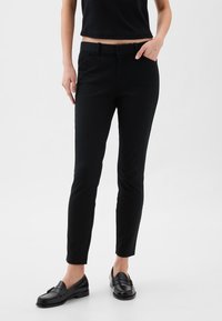 Fekete, testhezálló nadrág sima textúrával, övhurkokkal és oldalzsebekkel, fekete mokaszinokkal párosítva. Egyszerű, letisztult kialakítás.