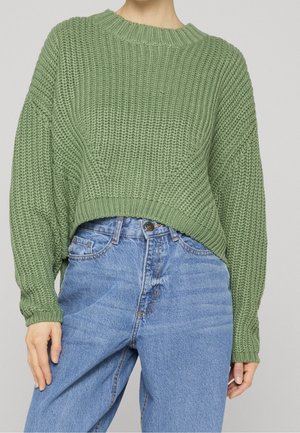 Mujer con un suéter de punto grueso verde claro con hombros caídos y vaqueros azules sueltos de talle alto sobre un fondo liso.