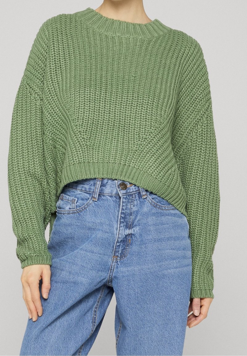 Femme portant un pull en grosse maille vert clair avec des épaules tombantes et un jean bleu ample taille haute sur un fond uni.