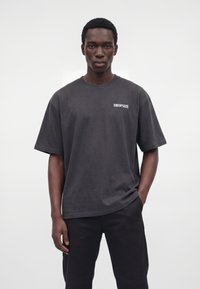 Jeune homme portant un T-shirt noir ample avec le texte « DROPSIZE » sur la poitrine et un pantalon noir, debout sur un fond blanc uni.