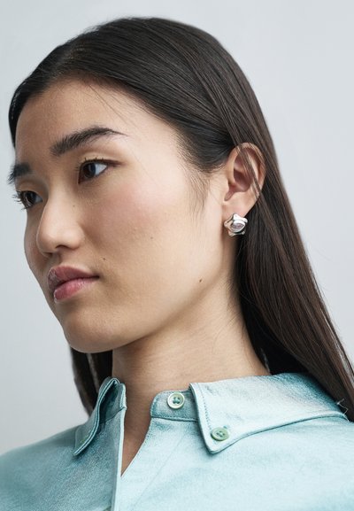 Boucles d'oreilles en argent avec un design arrondi et des accents texturés. Le modèle porte une chemise en satin bleu clair avec un col et des boutons verts.